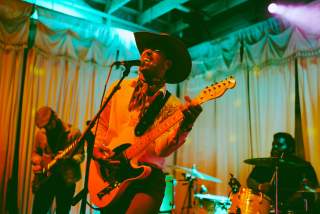 Musician in a cowboy hat singing into a mic while playing guitar with colorful lights surrounding him.