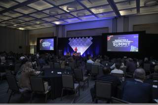 Woman standing on stage at DI Marketing & Communications Summit 2025, giving a welcome message to attendees in a ballroom.