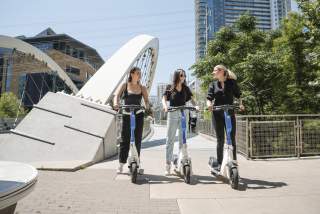 Three young women on electric scooters in front of downtown Austin's Butterfly Bridge.