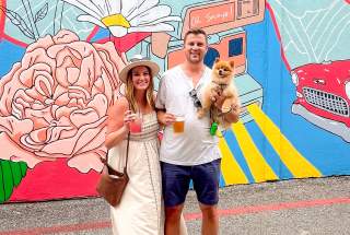 A couple stands in front of a vibrant mural, holding drinks, with a small Pomeranian dog nestled in the man's arms.