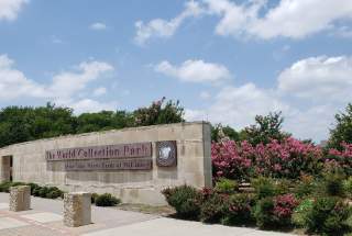 Sign for The World Collection Park, surrounded by blooming pink crape myrtle trees and greenery under a partly cloudy sky.