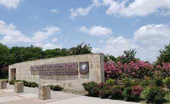 Sign for The World Collection Park, surrounded by blooming pink crape myrtle trees and greenery under a partly cloudy sky.