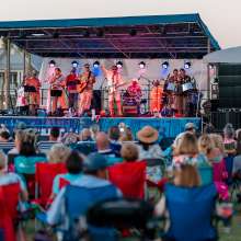 A band plays on a stage while a crowd of people in lawn chairs observes.
