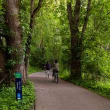 Two bikers riding on the Lansing River Trail surrounded by lush green plants