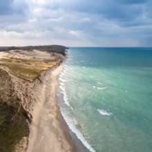 Aerial view of a lakeshore. The water on the right is a variety of light and dark blues. The dunes end in green vegetation and woods.