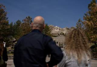A man and a woman, slightly out of focus in the foreground, look up at Mount Rushmore, in focus in the distance positioned between their heads.