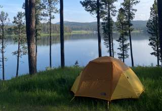 A yellow tent is set in the grass in the shadow of tall ponderosa pine trees under a blue sky overlooking a lake in the Black Hills.