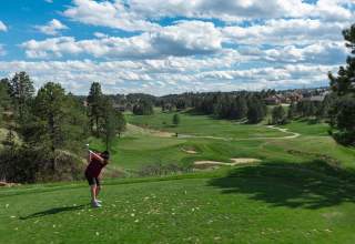 A man dressed in a black polo and black shorts tees off at the Golf Club at Red Rocks. Pine trees, a perfectly manicured course, bunkers, and a partly cloudy sky are visible in the distance.