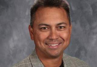 A formal headshot of Wayne Sullivan, dressed in a subtly checkered sport coat and black undershirt.