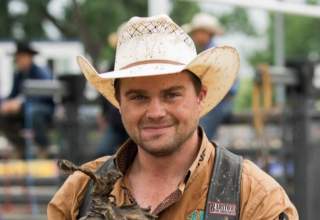 Shane, dressed in his rodeoing attire, holds a trophy of a cowboy on a bucking horse.