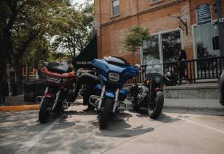 Three motorcycles, one red, one blue, and one black, are parked along the sidewalk, partially shaded by a tree, outside of Prairie Edge in Downtown Rapid City in mid afternoon.