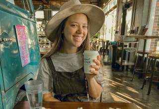 A 20-something girl in a wide brim hat smiles at the camera while holding a to-go cup of coffee in a well-lit coffeeshop. In front of her on the table are two waffles on plates, each covered in fruit and whipped cream.