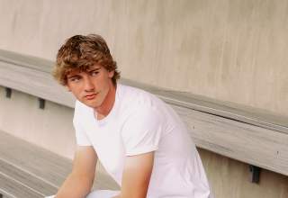 A senior portrait of Broeden Sales, pictured sitting in a baseball dugout in a white tshirt and jeans with his baseball cap, glove, and a baseball sitting next to him.