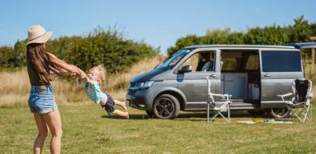 A woman swinging a toddler by his arms in front of a camper van.