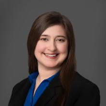 A young white woman with long brown hair smiles at the camera. She is wearing a bright blue button-down shirt and a black blazer in front of a solid gray background.
