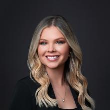 A young woman smiles at the camera. Her blonde hair is curled loosely over her shoulders. She is wearing a small silver pendant, a black top, and a black sweater on a black backdrop.