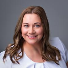 Amber Parsley smiles at the camera. She has shoulder-length brunette hair and light blue eyes. Her dress is either white or pale blue. The backdrop is solid gray.