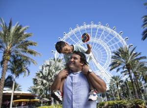 A boy on top of his dad's shoulders, while smiling and holding a lollipop, in front of The Wheel at ICON Park