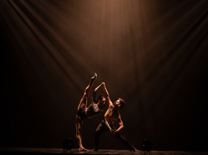 Two contemporary dancers perform an expressive lift under dramatic stage lighting.