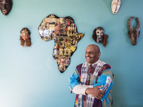 black man in traditional dress standing in front of a teal wall with a map of Africa
