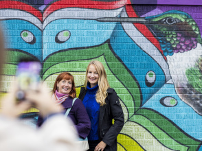 two women posing for a cell phone photo in front of a colorful mural of a bird