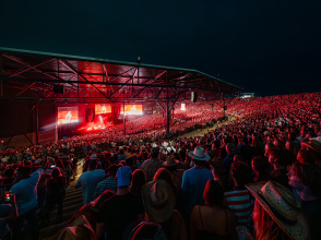An open-air concert stage with thousands of people watching a show illuminated in a red glow from the stage lights against the pitch black of the night sky.