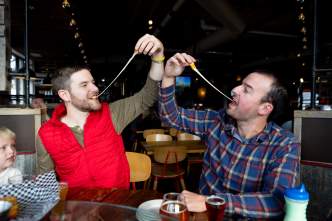 Two white men face each other while pulling strings of cheese curd from their mouth at Lone Girl Brewing