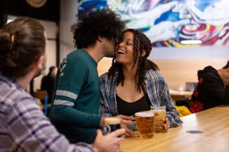 A Black man whispers into the ear of a Black woman at a bar at Working Draft Brewery while enjoying beers.
