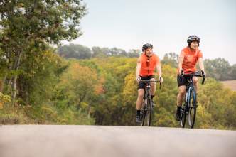 Two men biking up hill wearing bright orange shirts, helmets, and biking shorts. The trees in the background show leaves just beginning to change color for Fall.