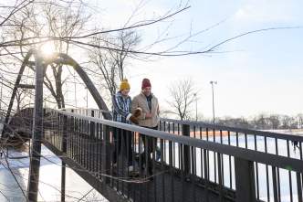 A couple walks across an arched bridge in Tenney Park during winter, surrounded by snow