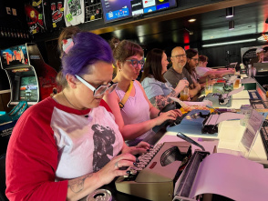 A group of people sit side by side at a bar counter, each using a typewriter. The person in the foreground has purple hair tied back with a scrunchie, glasses, and a red-and-white shirt with a graphic print. Others focus intently on their typewriters, with drinks and cans on the counter. Behind them, arcade machines, merchandise, and a TV screen are visible, creating a lively retro atmosphere.