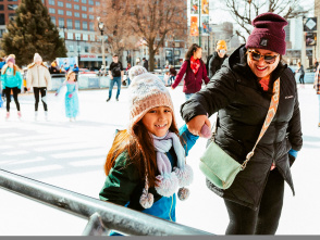 a little girl ice skating on a public ice rink while holding her mom's hand