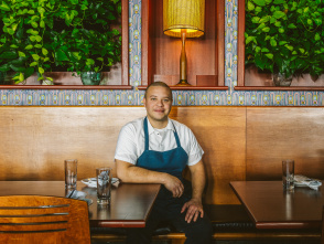 A chef wearing a white shirt and blue apron sits at a wooden booth inside a restaurant, smiling at the camera. The background features leafy green plants, patterned tile accents, and a warm lamp centered above him.