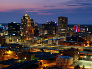 A twilight view of downtown Milwaukee, Wisconsin, featuring illuminated skyscrapers and city lights against a purple and blue sunset sky, with highways and streets visible in the foreground.