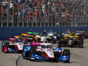 A dynamic shot of several IndyCars speeding along a track, led by a white, pink, and blue #60 car, with a crowd visible in the background.