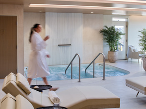 A tranquil spa lounge features cushioned reclining chairs, rolled towels, and a bubbling hydrotherapy pool beneath warm wood-paneled ceilings. A guest in a robe walks through the bright, modern space, creating a relaxing atmosphere focused on wellness and rejuvenation.