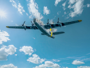 A historic four-engine bomber aircraft flies overhead against a bright blue sky dotted with white clouds. The polished silver plane is captured from below, highlighting its distinctive wingspan and vintage aviation design.