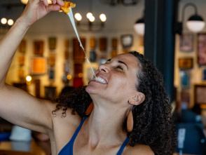 A smiling woman pulls apart a cheesy appetizer, creating a long stretch of melted cheese as she enjoys a meal inside a warmly lit restaurant. The background is softly blurred with artwork and lights visible.