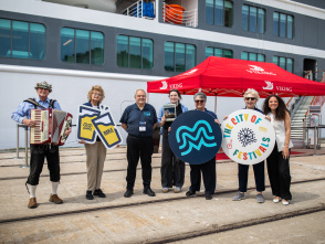 A welcoming committee with an accordion player stands before a cruise ship, holding Milwaukee-themed signs including an MKE logo and "City of Festivals" circular display, under a red Viking Cruises tent.