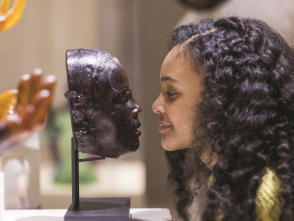 A young visitor leans in to closely examine a glass face sculpture displayed on a stand at a museum. The side profiles of the sculpture and the visitor align, creating a striking visual comparison in the gallery setting.