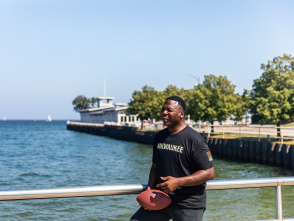 A man, LeRoy Butler, holding a football and smiling while leaning on a metal railing by a waterfront. Trees line the pathway behind him, and a building extends into the water in the background. The weather is clear and sunny.
