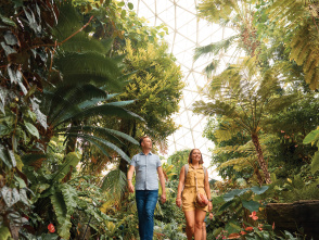 Two adults walk along a lush indoor garden path beneath a glass-domed ceiling, surrounded by tall palms, ferns and tropical plants inside a conservatory.