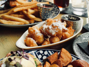 A cozy brunch spread featuring crispy potatoes, eggs Benedict, powdered sugar–dusted fritters with dipping sauce, and a sandwich with fries, all paired with iced tea and water on a sunlit table.