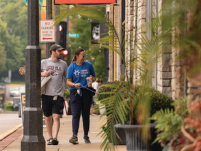 Two people stroll along a lively downtown sidewalk lined with local shops, hanging business signs, and colorful planters. Decorative lights and welcome banners add to the inviting streetscape as they explore the district on a warm day.