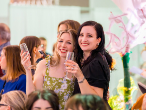 Two women smiling and posing together at a crowded indoor event, each holding a champagne flute, with floral decorations and other guests in the background.