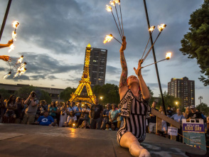 A fire performer in a striped costume kneels and raises flaming batons during a lively Bastille Days event in Milwaukee, with a crowd watching and a glowing Eiffel Tower replica lit up in the background.