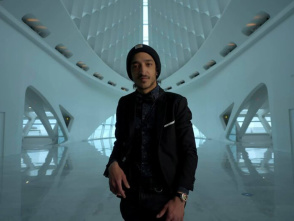 A man in a dark suit and beanie stands in the center of a large, futuristic, white architectural space with high arched ceilings and smooth reflective floors. The background features symmetrical structural elements and soft natural light coming from a tall window behind him.
