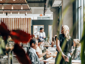 Diners enjoy a meal inside a bright, modern Milwaukee restaurant with large windows, warm lighting, and a relaxed, welcoming atmosphere as a chef engages with guests.