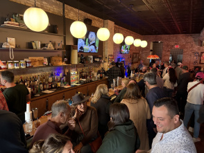 A crowded bar with warm lighting and exposed brick walls. People are gathered closely together, talking and drinking. A long bar counter is lined with bottles and glassware, and several round hanging lights illuminate the space. Two TVs mounted on the back wall display a show, and the overall atmosphere appears lively and social.