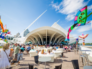 A wide angle shot of the Milwaukee Art Museum with festival goers sitting on the patio outside, with flags blowing in the wind against a beautiful blue sky.